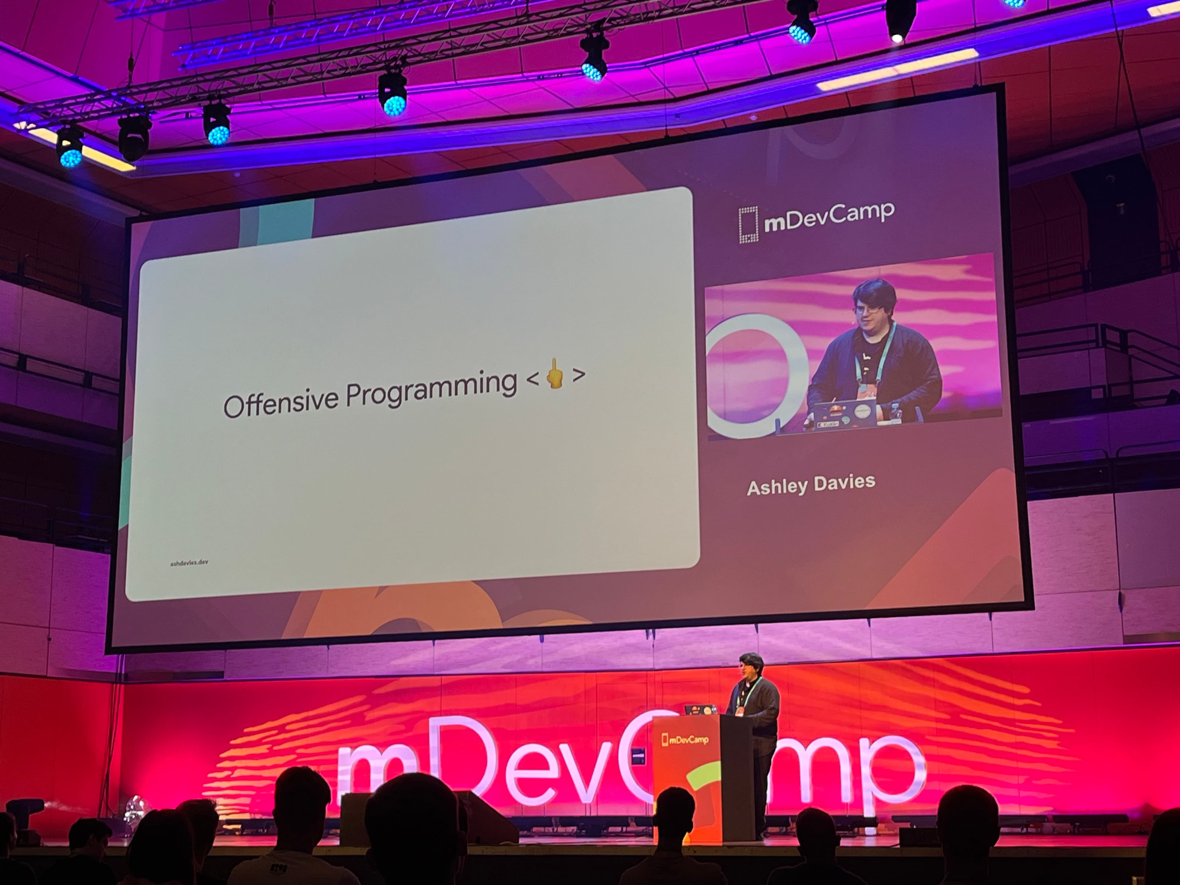Prezentujúci stojaci pod plátnom pri rečníckom pultíku prezentujúci na tému: "Offensive programming".