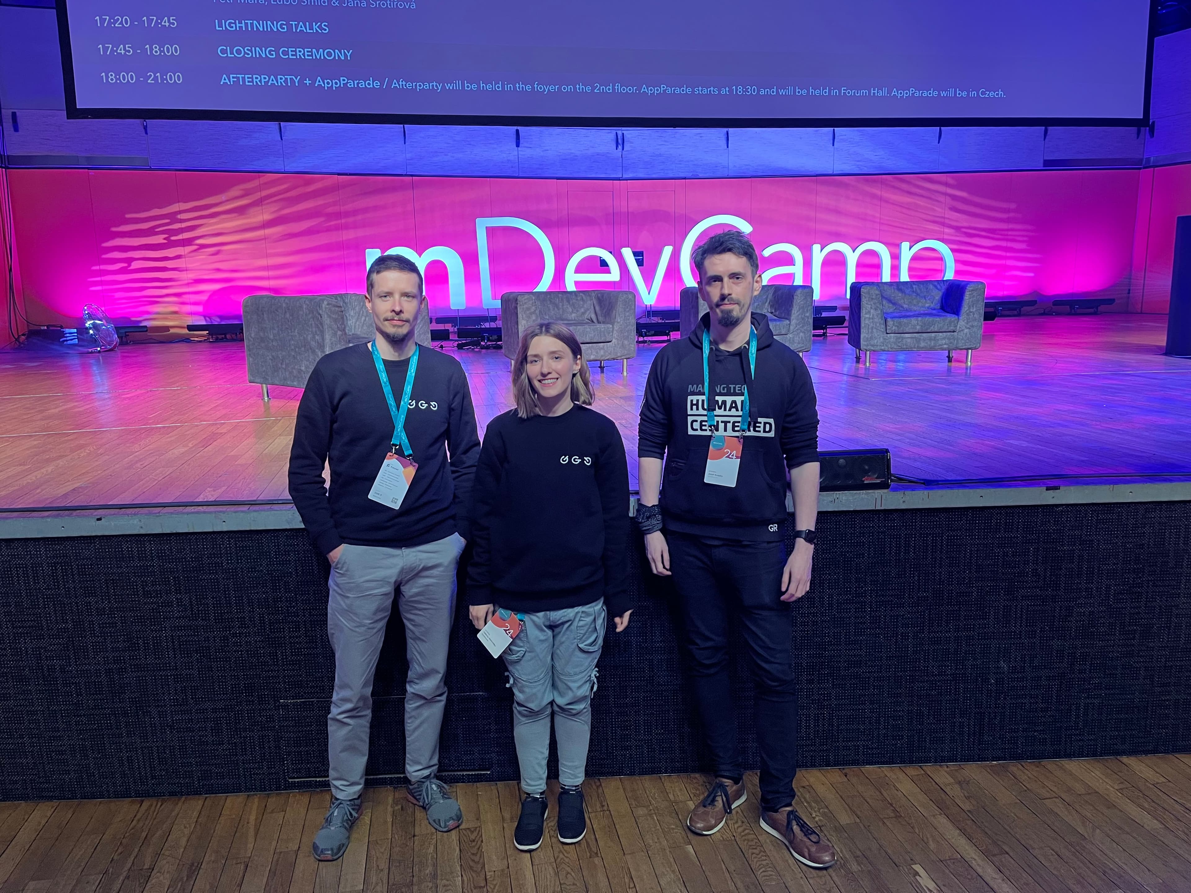 Tri osoby - dvaja muži a jedna žena, účastníci mDevCampu, stojaci pred veľkým nápisom mDevCamp.