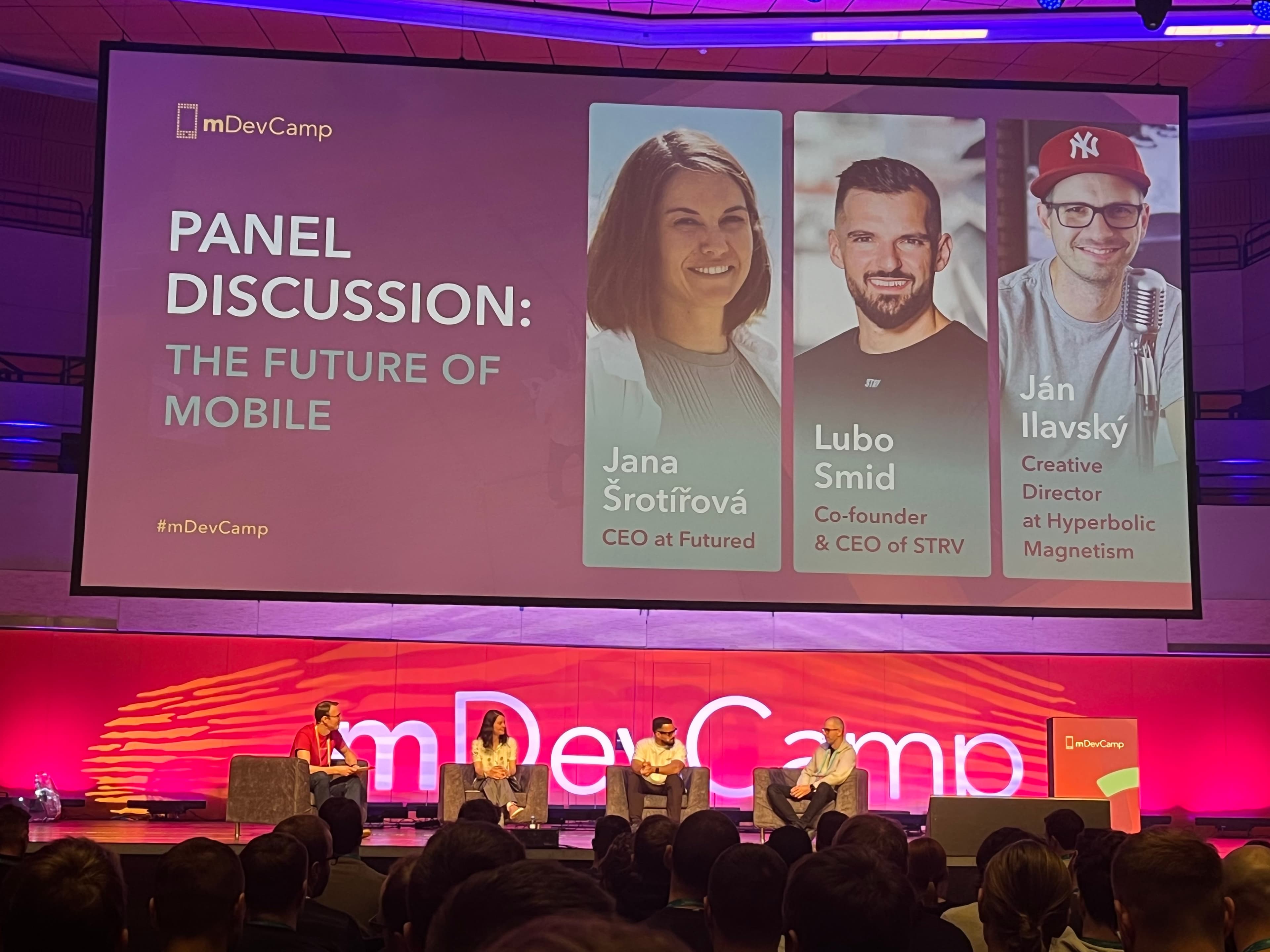 Plátno s nápisom "panelová diskusia" v rámci mDevCampu na tému: "The future of mobile", predstavujúca tri osoby.
