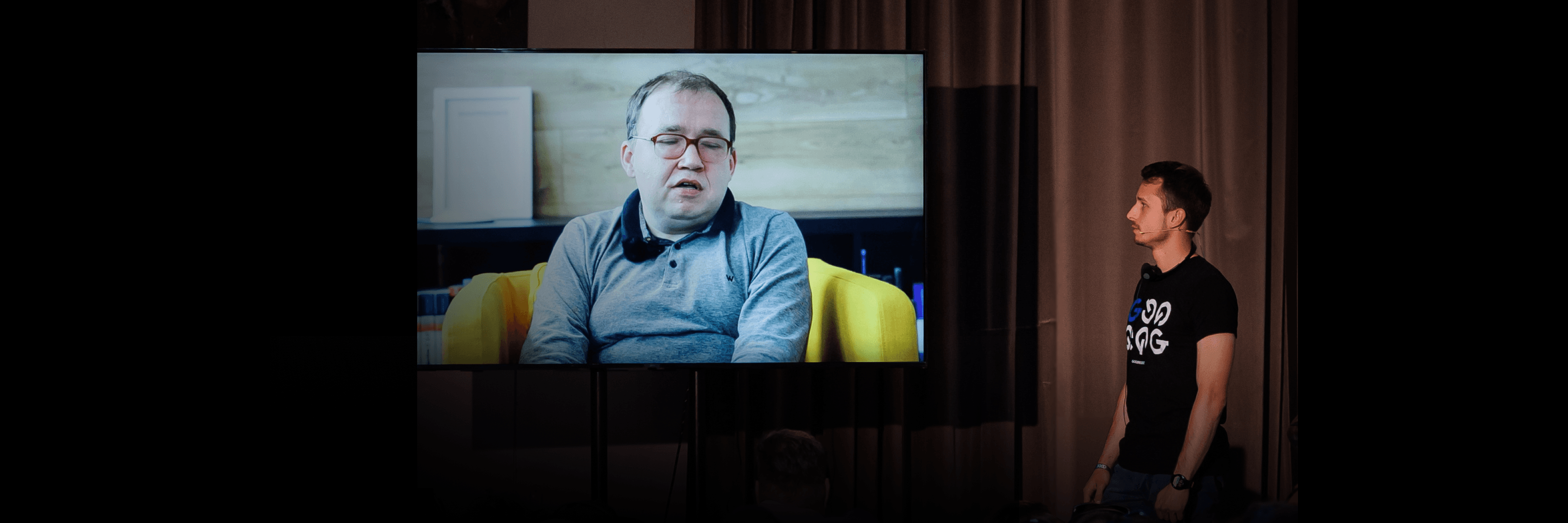 Obraz moderátora stojaceho na pódiu vedľa veľkej obrazovky, na ktorej sa zobrazuje video inej osoby.