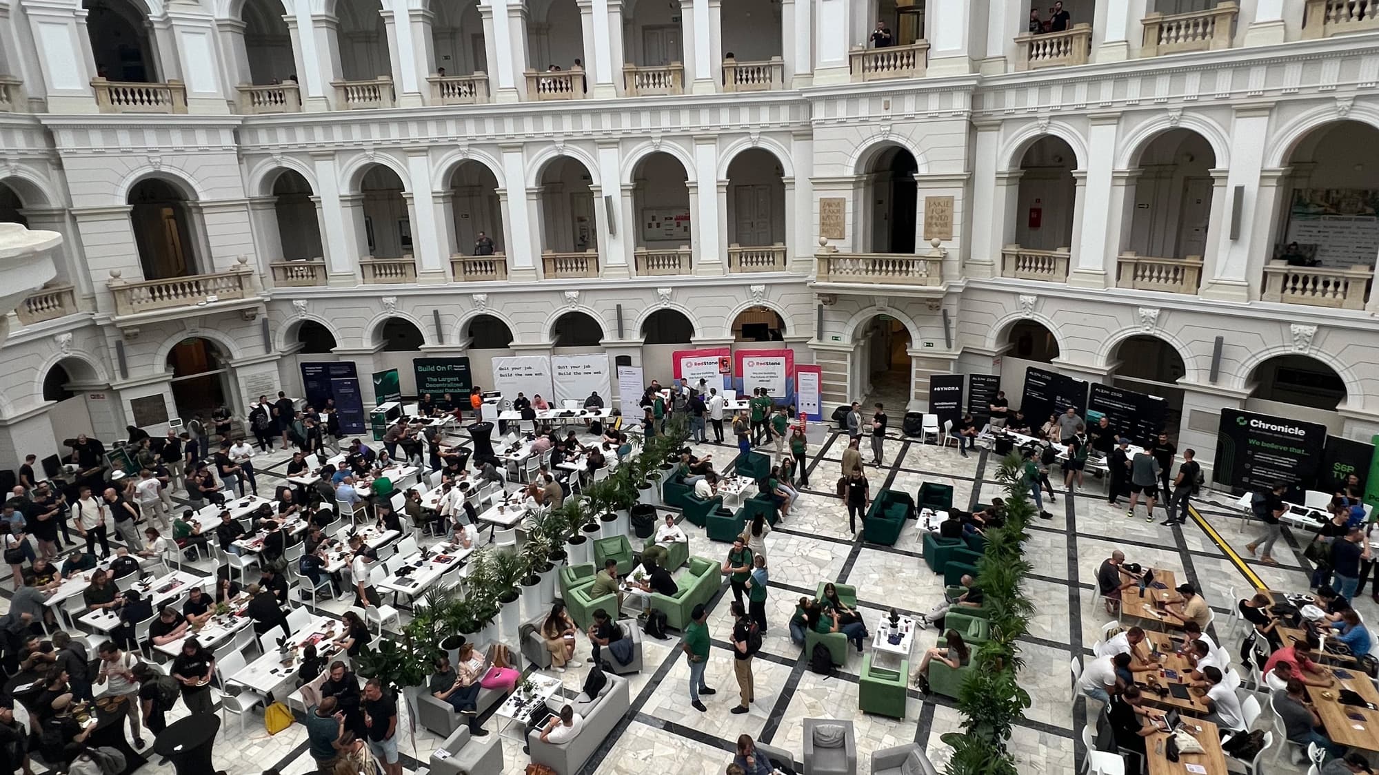 Konferenčná hala s oblúkmi a stĺpmi, kde ľudia sedia pri stoloch, pracujú na notebookoch a diskutujú. Po obvode sú stánky a bannery - odborné podujatie alebo konferencia.