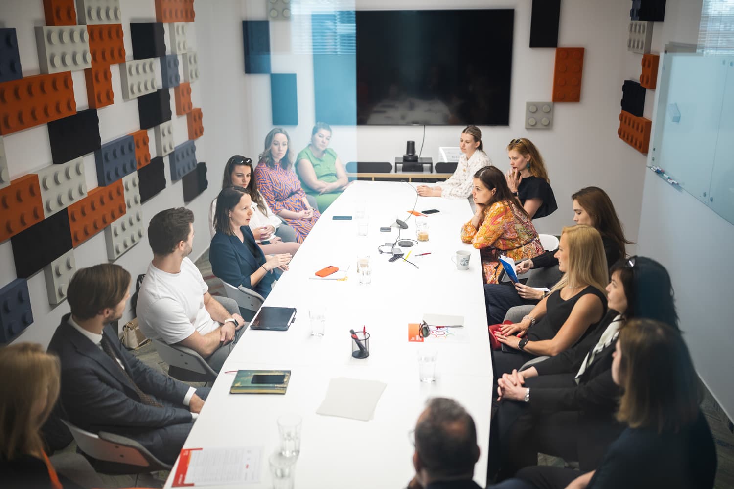 Skupina ľudí sedí okolo veľkého konferenčného stola v zasadacej miestnosti. Na stenách sú farebné akustické panely, vpredu je veľká obrazovka. Na stole sú poznámkové bloky, perá a poháre s vodou.